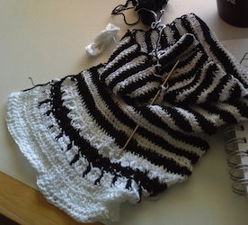 [IMAGE: photograph of a half-made striped black and white scarf. There is a crochet hook stuck through the scarf and balls of yarn nearby.]